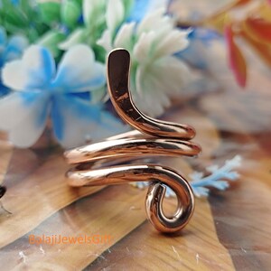 Pode incluir: Um anel de metal polido em tom de cobre com um design em espiral contínua que envolve o dedo. A banda tem uma superfície lisa e reflexiva e termina numa pequena curva arredondada na extremidade. O texto BalajiJewelsGift é visível.