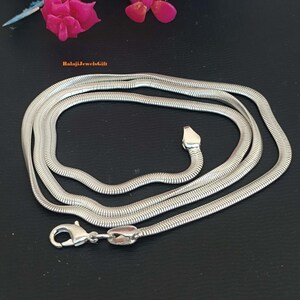 Könnte beinhalten: Eine silberfarbene Schlangenkette mit flachen, flexiblen Gliedern. Die Kette verfügt über einen Karabinerverschluss an einem Ende und eine kleine, spitz zulaufende Metalllasche am anderen Ende. Der Text BalajiJewelsGift ist in Orange sichtbar.