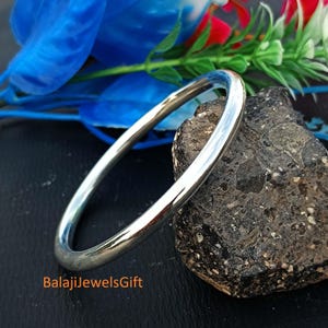 Peut inclure: Un bracelet jonc en métal argenté lisse et poli avec un profil arrondi. Le bracelet est placé à côté d'une pierre sombre, texturée et irrégulière. Le texte BalajiJewelsGift est imprimé en orange dans le coin inférieur gauche de l'image.