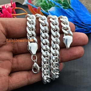 May include: A polished silver curb chain bracelet featuring thick, interlocking flat links and a secure lobster claw clasp. The metal surface is highly reflective and smooth. The text BalajiJewelsGift is printed in orange on the silver surface.