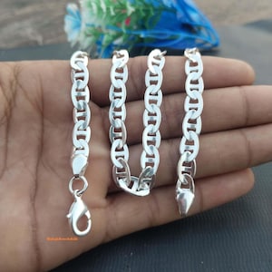 Puede incluir: Una pulsera de cadena en tono plateado con eslabones ovalados que presentan una barra horizontal central, conocida como cadena de estilo marinero o ancla. Incluye un cierre de mosquetón en un extremo y un acabado pulido en el metal.