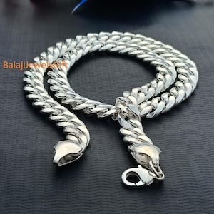 May include: A polished silver-toned curb chain necklace featuring thick, interlocking flat links and a secure lobster claw clasp. The metal surface reflects light, highlighting the uniform, rounded shape of each individual link in the chain.