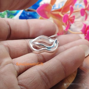 May include: A polished silver ring featuring a continuous, fluid, wavy design that creates an open, layered appearance. The band consists of smooth, interconnected metallic curves that wrap around the finger, displaying the text BalajJewelsGift.