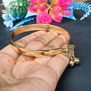May include: A polished gold-tone metal bangle bracelet featuring a smooth, rounded band and a small charm attachment with two tiny spherical bells hanging from a circular loop. The text BalajiJewelsGift is printed in orange on the lower left side.