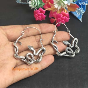 Peut inclure: Une paire de créoles argentées présentant un motif de serpent texturé qui s'enroule autour du cadre circulaire. Le métal a une finition oxydée pour souligner les écailles du serpent. Le texte BalajiJewelsGift est visible en orange.