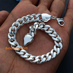 Könnte beinhalten: Ein glänzendes Panzerkettenarmband in Silberoptik mit dicken, ineinandergreifenden flachen Gliedern und einem sicheren Karabinerverschluss. Die Metalloberfläche hat ein hochglänzendes Finish und der Text BalajJewelsGift ist in Orange zu sehen.