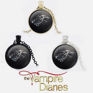 Könnte beinhalten: Drei Anhänger-Halsketten mit einem Wolfskopf-Design. Die runden Anhänger sind schwarz mit silberfarbenen Wolfsköpfen. Die Ketten und Fassungen sind goldfarben, schwarz und silberfarben. Der Text "the Vampire Diaries" ist unten.