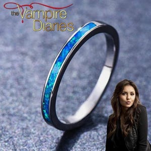Op de afbeelding: Een zilveren ring met een band van levendig blauwe opaal. De ring staat tegen een getextureerde grijze achtergrond. De tekst "The Vampire Diaries" staat bovenaan.