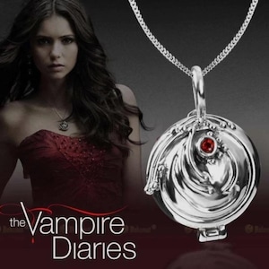 Puede incluir: Un collar con colgante plateado con un diseño redondo y ornamentado y una pequeña gema roja. El collar está en una cadena plateada. El texto "the Vampire Diaries" es visible en la esquina inferior izquierda.