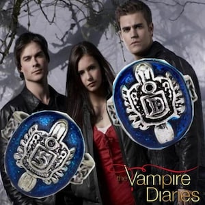 May include: Silver ring with a blue enamel face, displaying a crest design and the letter "D". The ring is from "The Vampire Diaries". The image shows the ring in front of three people.