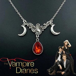 Puede incluir: Collar plateado con un colgante central de gema roja en forma de lágrima. El collar tiene elementos decorativos de filigrana y dijes de media luna. El texto "The Vampire Diaries" es visible en la esquina inferior izquierda.