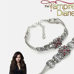 Könnte beinhalten: Silberfarbenes Armband mit floralem Design und roten Akzenten. Das Armband hat detaillierte Muster und einen Kettenverschluss. Der Text "The Vampire Diaries" ist oben rechts sichtbar.