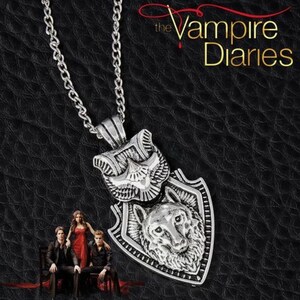 Puede incluir: Collar con colgante plateado con un diseño de lobo dentro de un escudo, con un emblema de ave encima. La cadena es de eslabones. Las palabras "The Vampire Diaries" están en letras doradas.