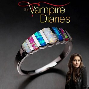 May include: Silver ring with a series of colorful opal stones. The ring features alternating sections of white and vibrant colors, including pink, blue, and green. The text "The Vampire Diaries" is displayed at the top.