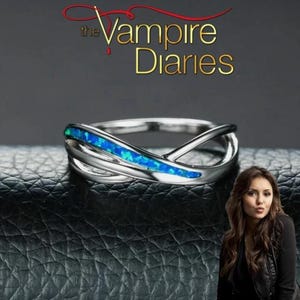 Op de afbeelding: Zilverkleurige ring met een blauwe opaal inleg. De ring heeft een verstrengeld ontwerp met de gouden tekst "The Vampire Diaries" erboven. De ring wordt gepresenteerd op een zwarte, getextureerde ondergrond.