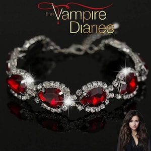 May include: A silver bracelet with large, teardrop-shaped red gemstones, each surrounded by smaller, clear crystals. The bracelet is set against a black background, with the text "The Vampire Diaries" in gold.