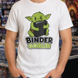 May include: White t-shirt featuring a green goblin character holding a black binder. The goblin has large pointed ears and is wearing a brown loincloth. The text "BINDER GOBLIN" is printed below the goblin in black and green.