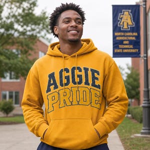May include: A mustard yellow hoodie with the text "AGGIE PRIDE" in navy blue and gold lettering. The person is standing in front of a university banner. The hoodie has a drawstring hood and a front pocket.