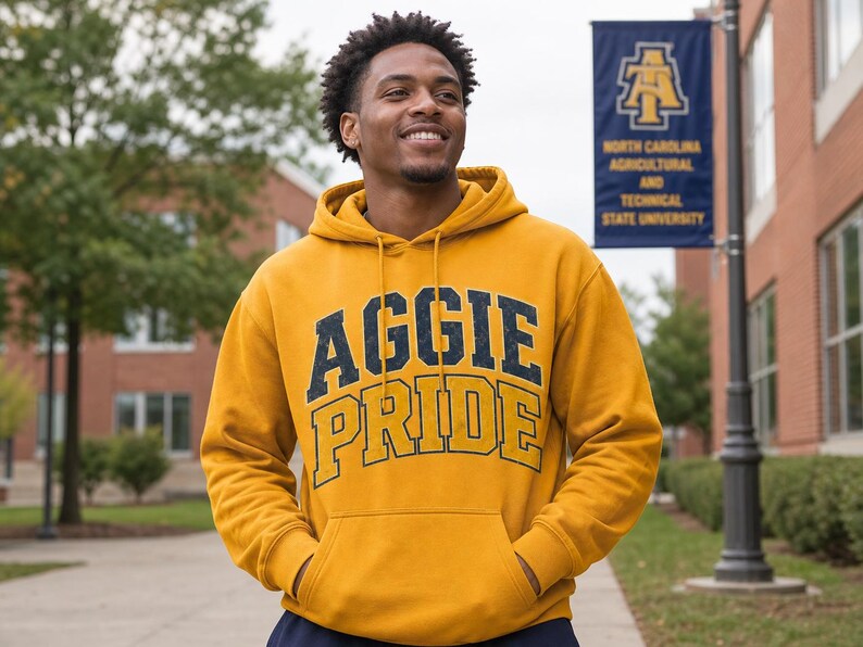 May include: A mustard yellow hoodie with the text "AGGIE PRIDE" in navy blue and gold lettering. The person is standing in front of a university banner. The hoodie has a drawstring hood and a front pocket.