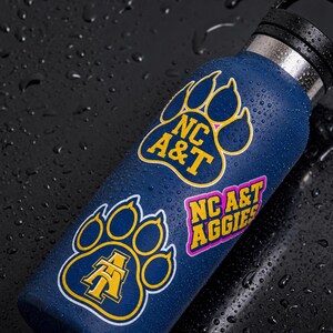 May include: A navy blue water bottle with black lid, adorned with stickers. The stickers feature the text "NC A&T" and "NC A&T AGGIES" in yellow and pink, set within paw print designs. The bottle is covered in water droplets.