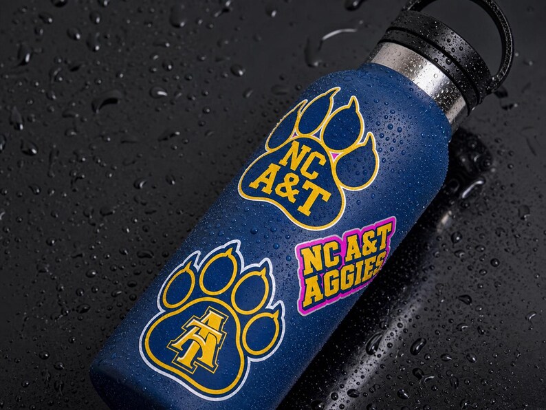 May include: A navy blue water bottle with black lid, adorned with stickers. The stickers feature the text "NC A&T" and "NC A&T AGGIES" in yellow and pink, set within paw print designs. The bottle is covered in water droplets.