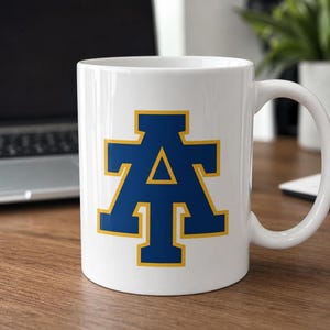 May include: White ceramic mug featuring a blue letter "A" with a gold outline. The mug has a curved handle and is set on a wooden surface. The design is centered on the mug's front.