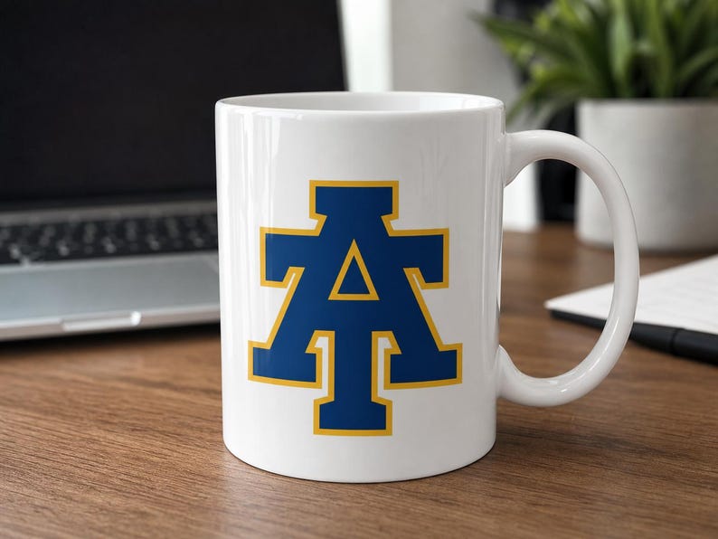 May include: White ceramic mug featuring a blue letter "A" with a gold outline. The mug has a curved handle and is set on a wooden surface. The design is centered on the mug's front.