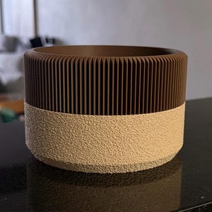 May include: A cylindrical brown and beige planter. The top section is a dark brown with vertical ribbed detailing. The bottom section is a lighter beige with a textured, speckled surface. The planter sits on a black surface.