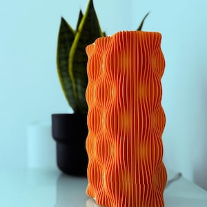 May include: An orange, wavy-patterned lamp with a vertical ribbed design. The lamp is cylindrical and sits on a white surface. A green plant in a black pot is in the background.