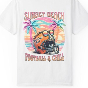 Peut inclure: Un t-shirt blanc avec un motif représentant un casque de football américain orange portant des lunettes de soleil, devant une plage au coucher du soleil avec des palmiers. Le texte indique SUNSET BEACH en haut et FOOTBALL & CHILL en bas.