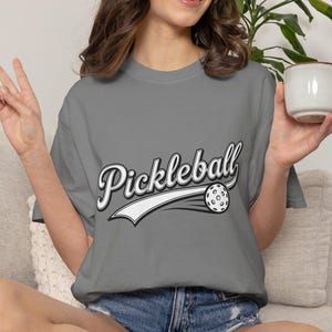 Peut inclure: Un t-shirt gris à manches courtes avec le mot Pickleball écrit en lettres blanches avec un contour noir. Sous le texte se trouve un graphique représentant une balle de pickleball blanche avec des trous, accompagnée de lignes de mouvement.