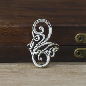 May include: A silver ring with an ornate, swirling design. The ring features a central, elongated loop with smaller, decorative curls and flowing lines. The ring is set against a dark wooden background.