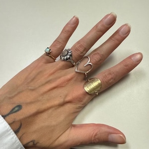 Puede incluir: Una mano que muestra cinco anillos. Los anillos incluyen un anillo de oro con un pequeño ópalo, un anillo de plata con forma de girasol, un anillo de plata en forma de corazón y un anillo de oro texturizado. La mano tiene un tatuaje en la muñeca.