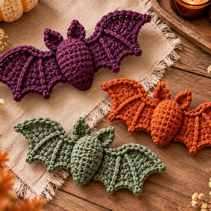 Peut inclure: Trois chauves-souris crochetées en violet, orange et vert. Chaque chauve-souris présente un corps texturé et rembourré avec deux oreilles pointues et de larges ailes plates avec des détails cousus visibles. Elles sont disposées sur un tissu clair.