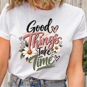 May include: A white short-sleeved t-shirt featuring a graphic design with the text Good Things Take Time in black, pink, and olive green script and block lettering, accented by white daisy flowers, green leaves, and small black heart outlines.
