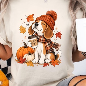 May include: A cream-coloured t-shirt featuring a graphic of a beagle puppy wearing an orange knit hat and a plaid scarf. The puppy holds a coffee cup labelled Coffee Lover. The design includes two pumpkins and scattered autumn leaves in orange and red.