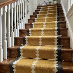 May include: A staircase with wooden steps and a runner rug. The runner is mustard yellow with a geometric pattern in white and gray. Brass rods secure the runner to the steps. White balusters and a wooden handrail are on the left.