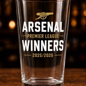 May include: A clear pint glass featuring the text "ARSENAL PREMIER LEAGUE WINNERS 2025/2026" in white and gold. The Arsenal logo is at the top. The glass is set on a wooden surface.