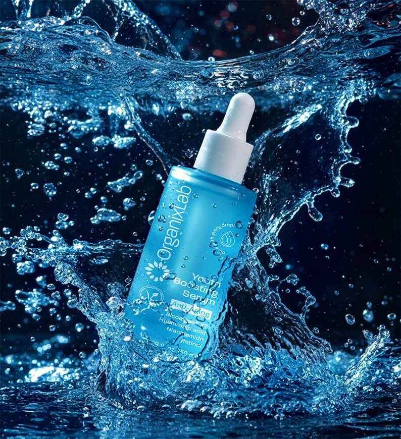 May include: A blue OrganixLab Youth Boosting Serum bottle with a white dropper is surrounded by splashing water. The bottle has white text and a flower logo. The serum contains ingredients like Hyaluronic Acid and Retinol.