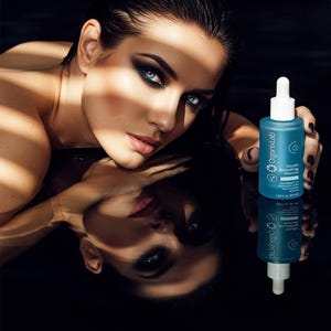 May include: A woman with dark hair and dramatic eye makeup holds a teal bottle of OrganixLab Youth Boosting Serum. The bottle has a white dropper and white text. The image is reflected in a dark surface.