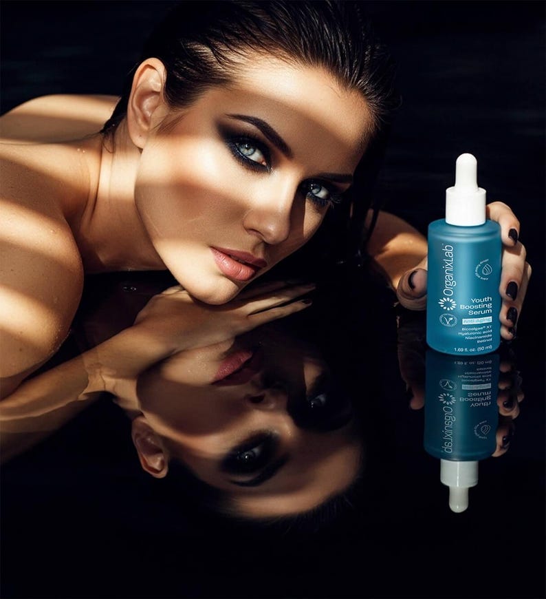 May include: A woman with dark hair and dramatic eye makeup holds a teal bottle of OrganixLab Youth Boosting Serum. The bottle has a white dropper and white text. The image is reflected in a dark surface.