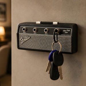 May include: A black Fender Mini Twin Amp key holder mounted on a wall. The key holder has three input jacks, a handle, and a silver and black speaker grill design. A set of keys hangs from a carabiner attached to the key holder.