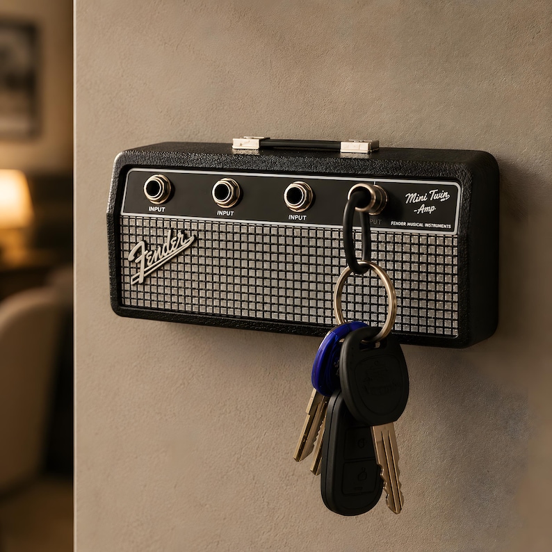 May include: A black Fender Mini Twin Amp key holder mounted on a wall. The key holder has three input jacks, a handle, and a silver and black speaker grill design. A set of keys hangs from a carabiner attached to the key holder.