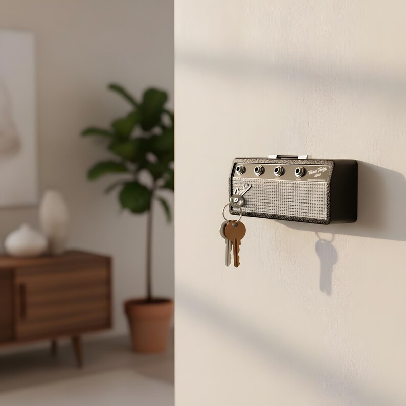 May include: A black key holder shaped like a vintage amplifier, mounted on a white wall. The key holder has silver-colored knobs and a speaker grill design. A set of keys hangs from the key holder.