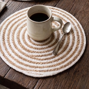 Op de afbeelding: Ronde placemats met een spiraalpatroon van afwisselend natuurlijk bruin jutekoord en wit katoenen koord. De cirkelvormige matten hebben een getextureerde, gevlochten constructie en een plat profiel, ontworpen als stabiele basis voor servies.