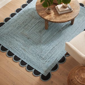 Puede incluir: Una alfombra rectangular que presenta un centro tejido en color azul claro con una superficie texturizada y un borde festoneado decorativo compuesto por elementos circulares tejidos en negro. La alfombra está hecha de fibra trenzada gruesa.