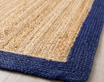 Puede incluir: Una alfombra rectangular con un centro de fibras tejidas en color beige natural y un borde grueso trenzado en azul oscuro. La textura es rugosa y uniforme, mostrando un primer plano detallado del material tejido y del borde de la esquina.