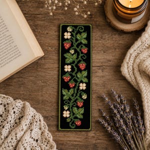 Può includere: Un segnalibro rettangolare con un motivo a punto croce di fragole rosse, foglie verdi e fiori bianchi su sfondo nero. Il design è orientato verticalmente con un motivo botanico ripetitivo di viticci e frutti.