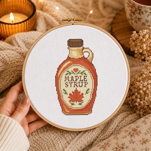 May include: A circular wooden embroidery hoop containing a cross-stitch design of a maple syrup bottle. The bottle is amber-coloured with a brown cap, a handle, and a label featuring the text MAPLE SYRUP above a red maple leaf graphic.