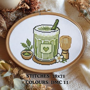 Puede incluir: Bordado de punto de cruz en un aro de madera que representa un matcha latte verde con un corazón, un batidor y una pequeña planta. El texto en la parte inferior dice: STITCHES: 78x71, COLOURS: DMC 11. El diseño está sobre un fondo blanco.